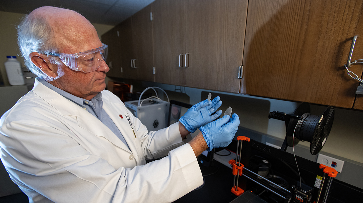 A man wearing a white lab coat and blue gloves holds a small medical device in a laboratory.