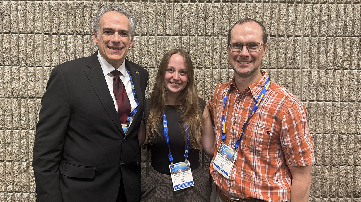Two men stand with a young woman, all wearing name badges, against an tan stone wall.