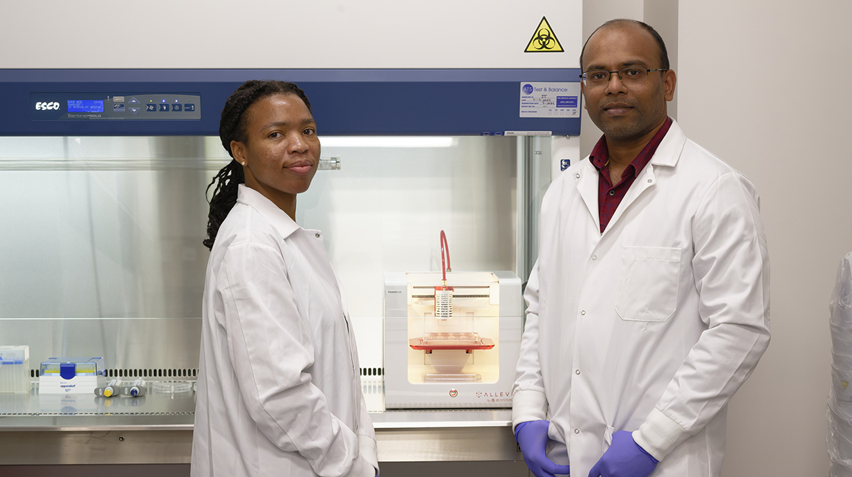 A woman and a man wearing white lab coats stand in front of a vented hood in a laboratory.
