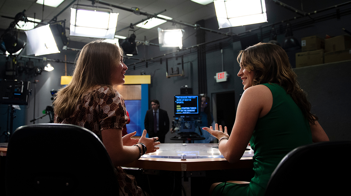 Two young women sitting at a desk chat in front of lights, TV cameras and a stage crew.
