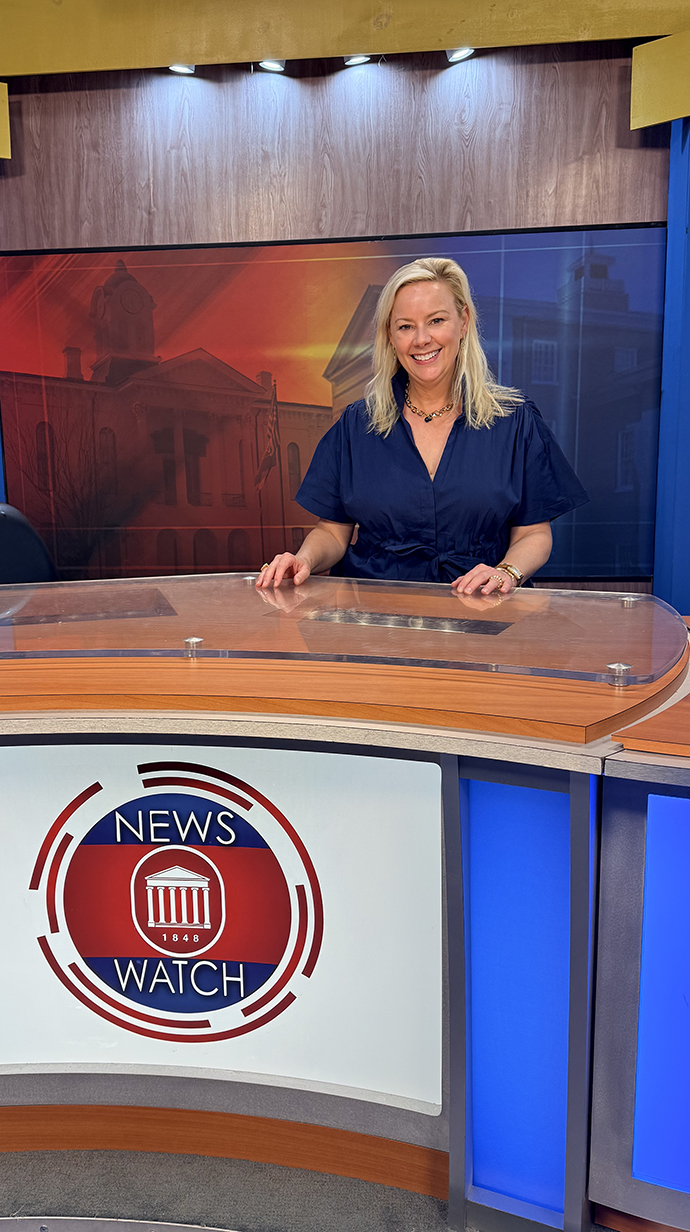 A woman sits at a desk on a TV news set.