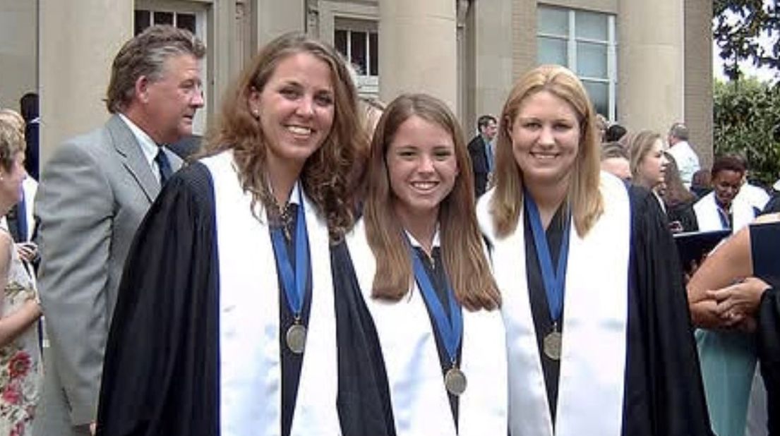 Three young women wearing graduation robes gather outside a large building with stone columns.