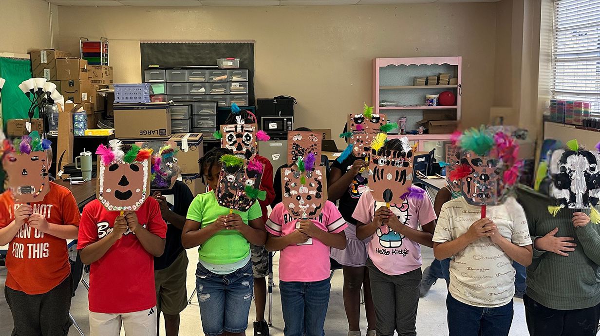 A line of children hold painted masks over their faces in a classroom.