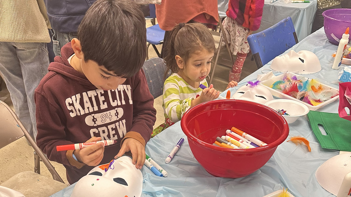 A young boy and girl paint white masks on a table in a classroom.