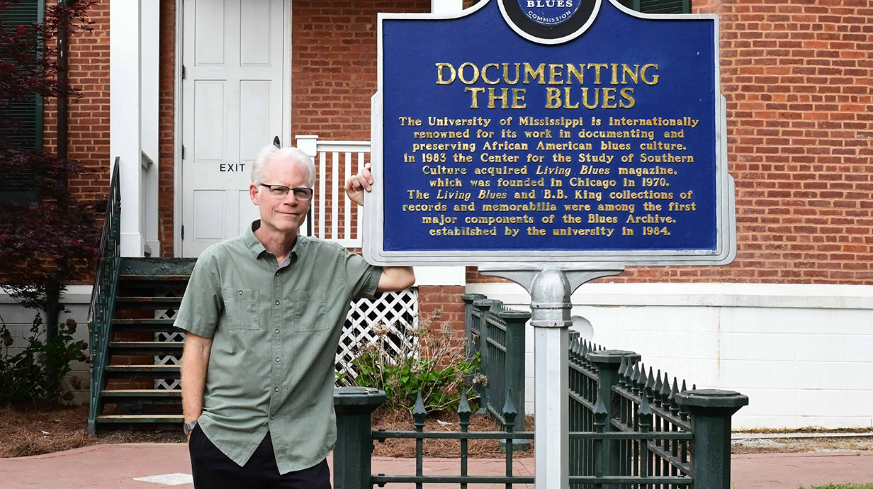 A man wearing a casual gray shirt stands next to a historical marker reading 'Documenting the Blues' outside a brick building with a high front porch.
