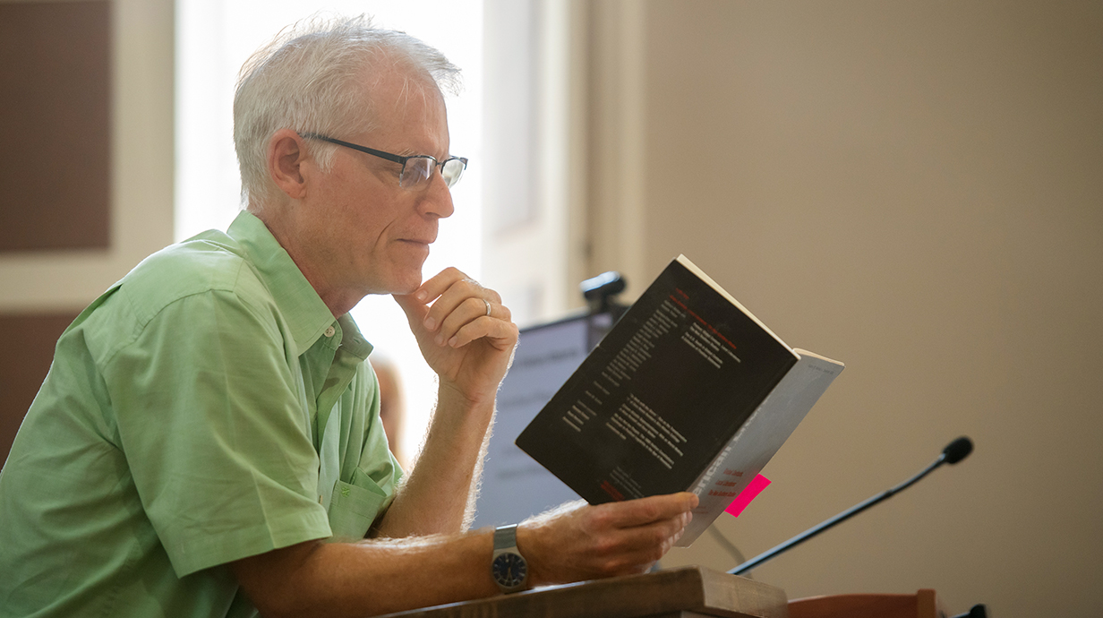 A man wearing glasses reads from a book at a lectern.
