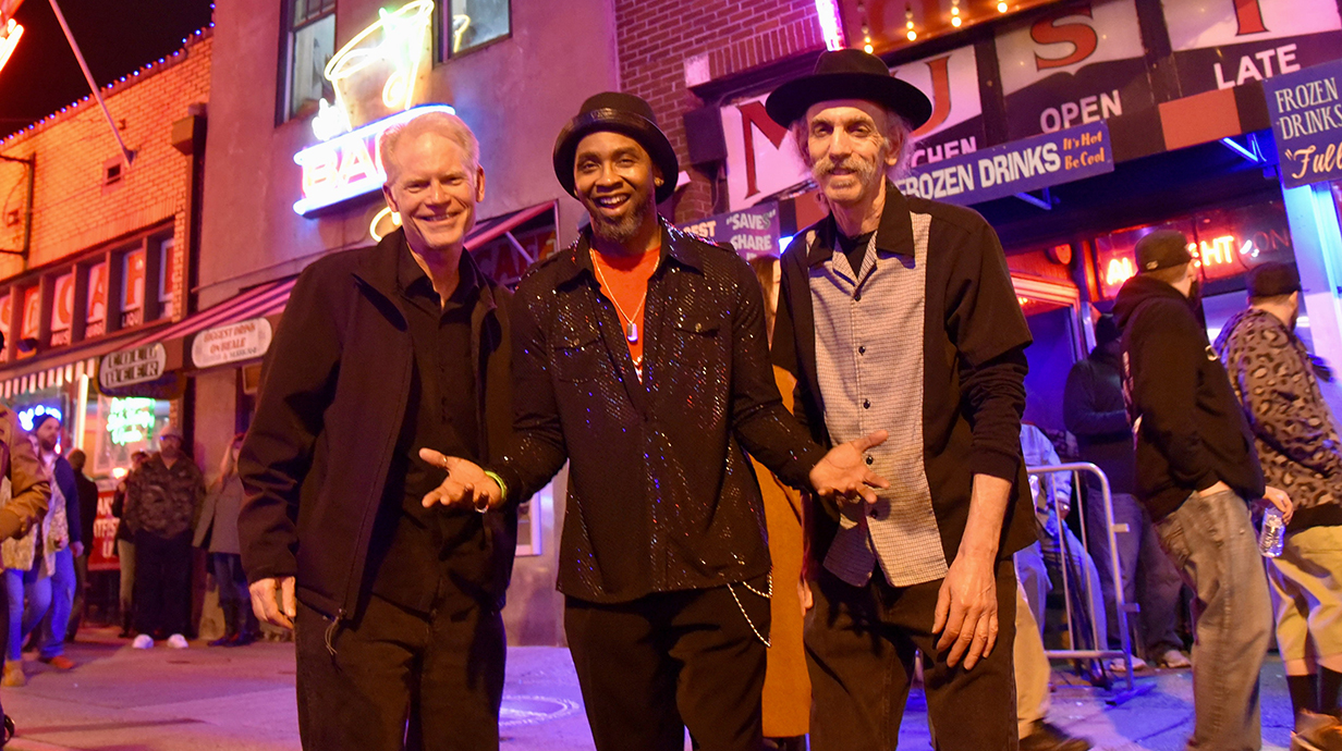 Three men stand on a brightly lit street at night.