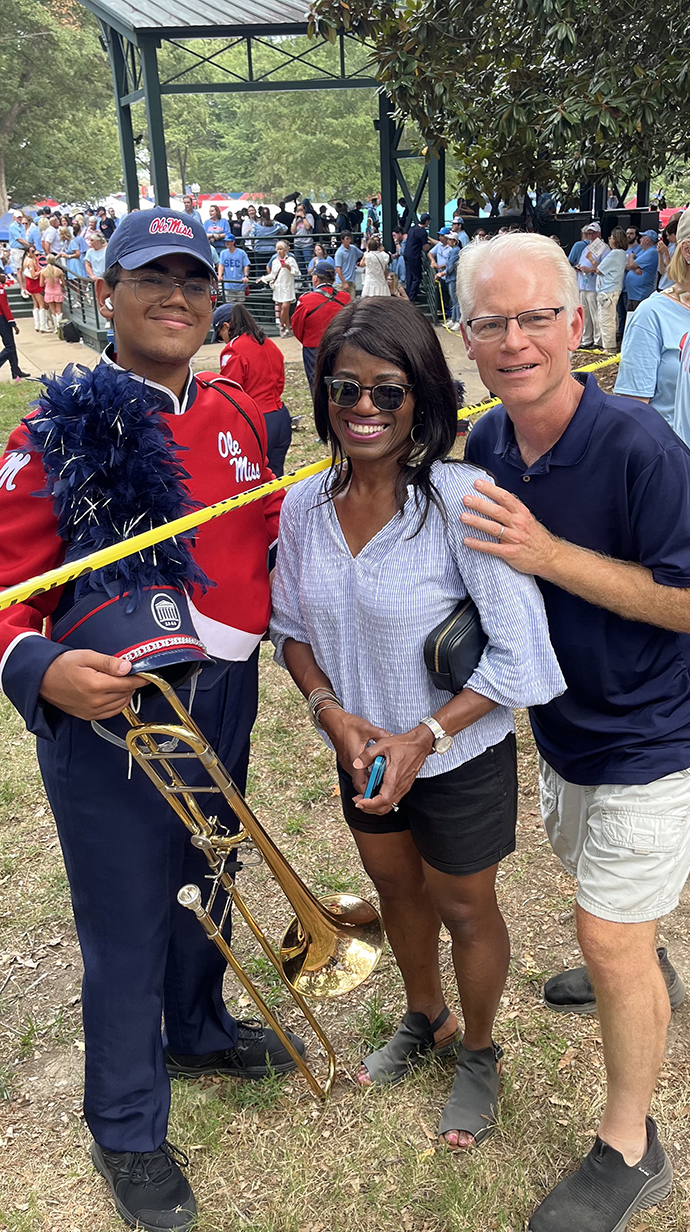 A man and woman stand with a young man wearing a band uniform in a wooded park.