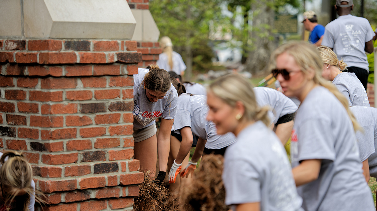 A group of young people spreads pine straw around brick columns in an outdoor courtyard.