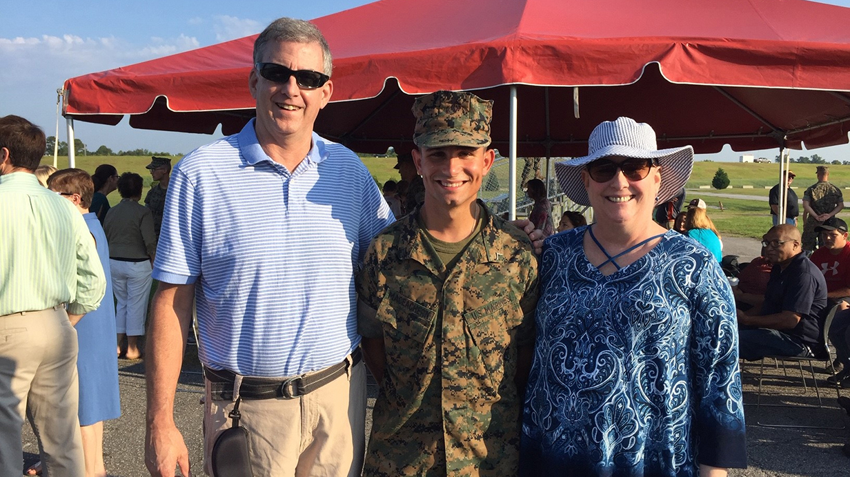 A woman stands with a man and a young man in a combat uniform in front of a pop-up tent on a sunny day.