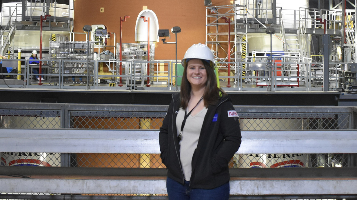 A woman wearing a white hardhat stands on front of a giant rocket being assembled at the Kennedy Space Center.