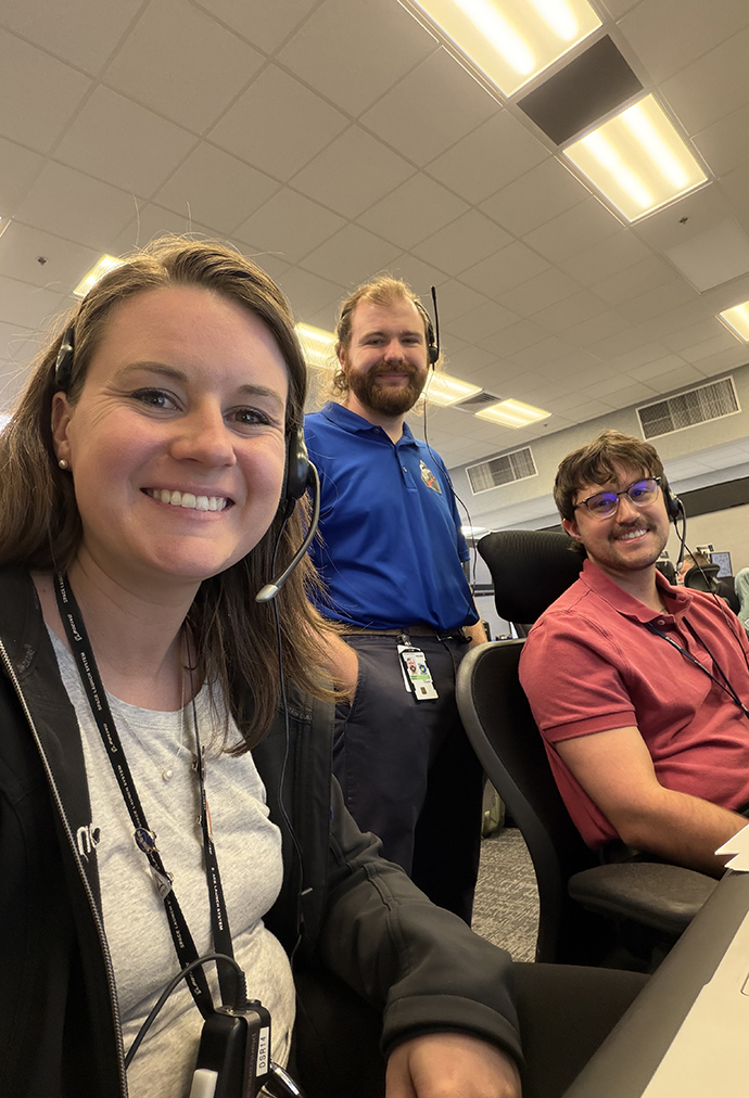 A woman and two men monitor computer terminals in a control room.