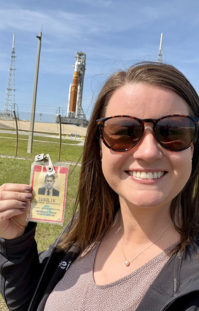 A woman standing in front of a large rocket holds up a vintage ID badge.