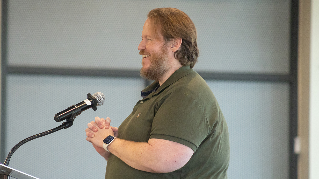 A man wearing an olive polo shirt speaks into a microphone in an auditorium.
