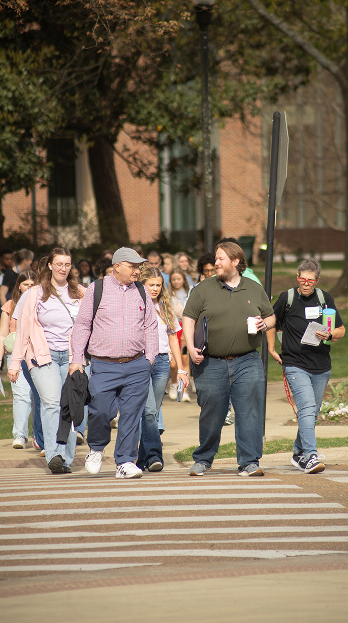 A large group of people walks through an intersection on a tree-lined street.