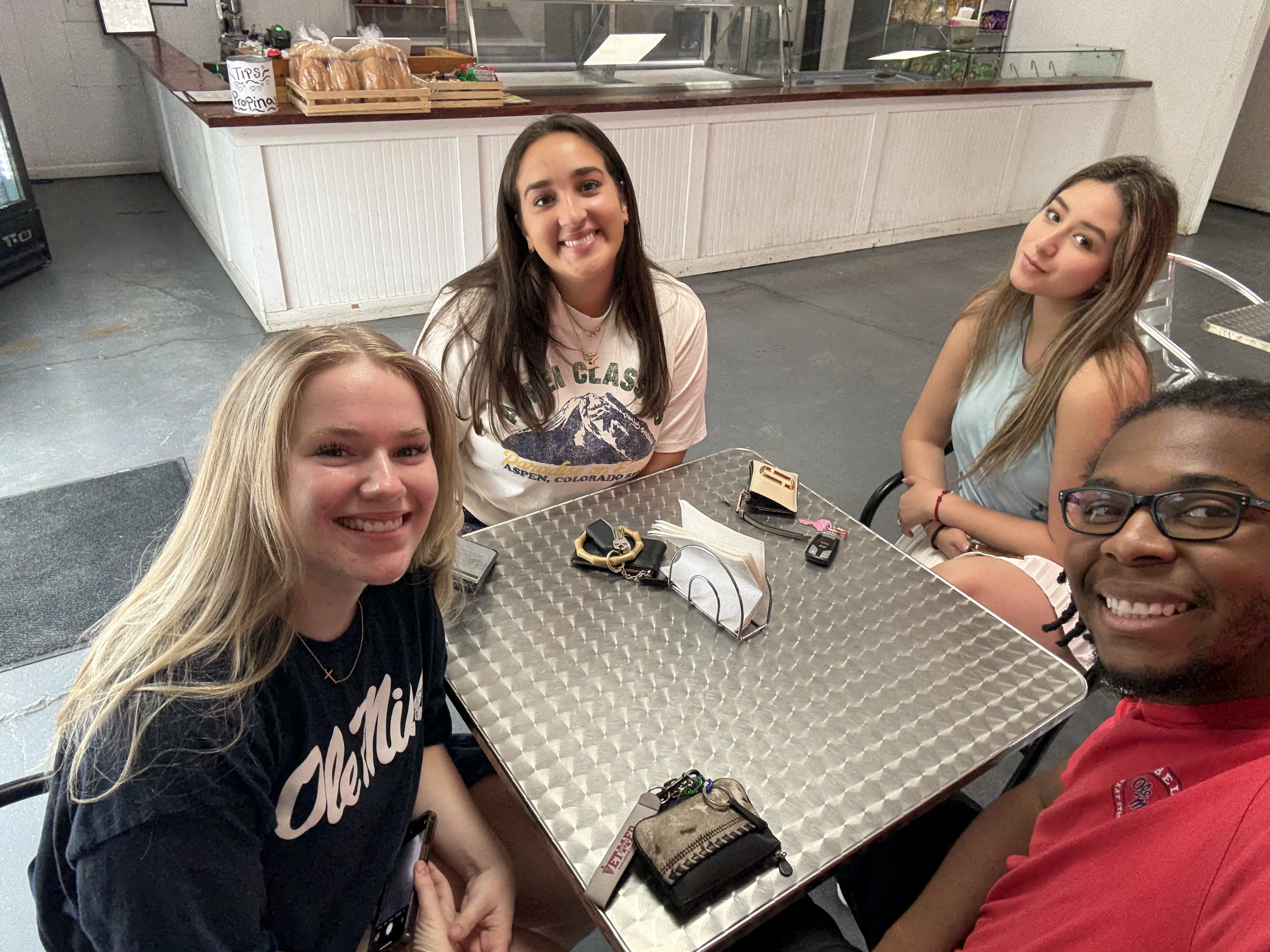 A young man and three young women seated around a small square table, look up and smile for the camera.