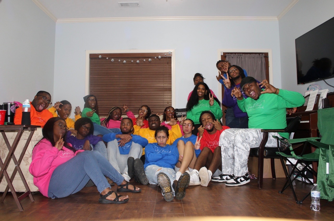 A large group of young people, seated on the floor and in a chair, pose for a fun group photo.