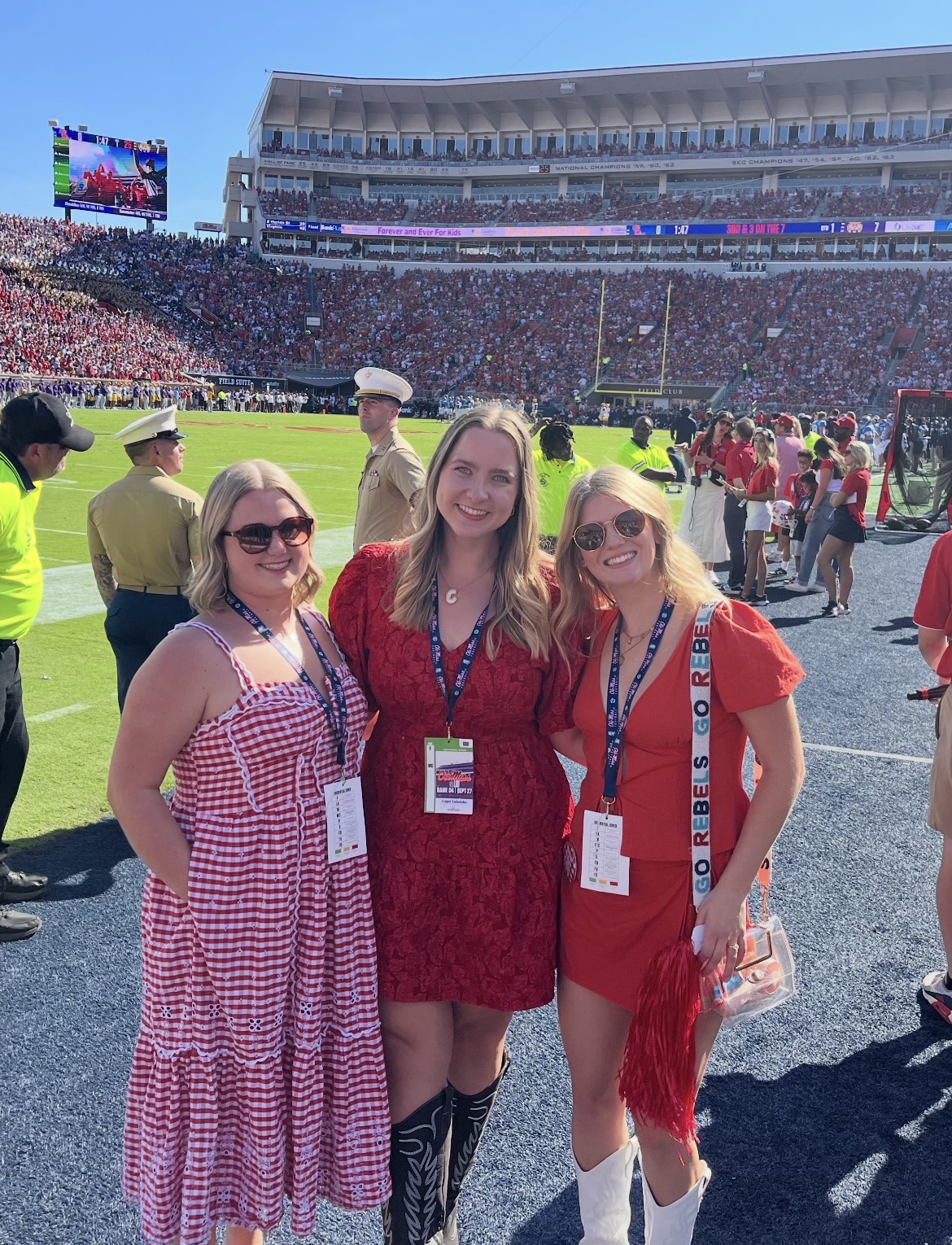 Three young women wearing ID badges on lanyards stand on the sideline of a football field before a game.