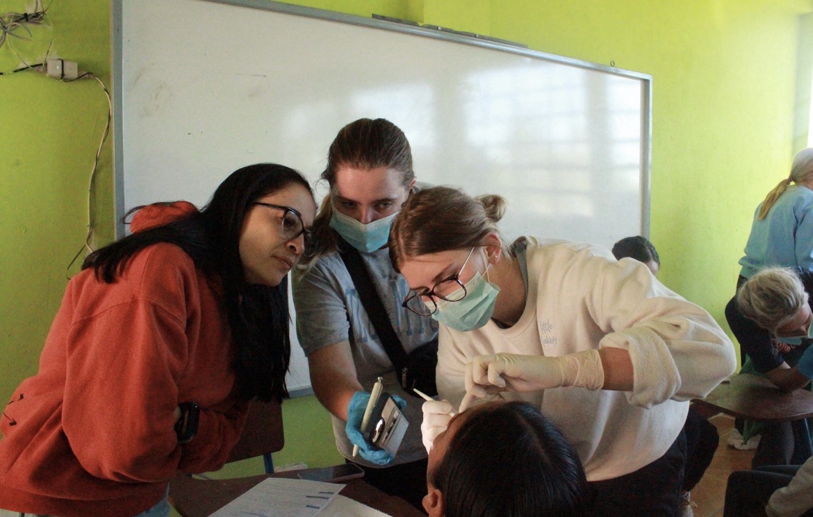 Two women watch while another woman performs an oral exam on a dental patient in a chair.