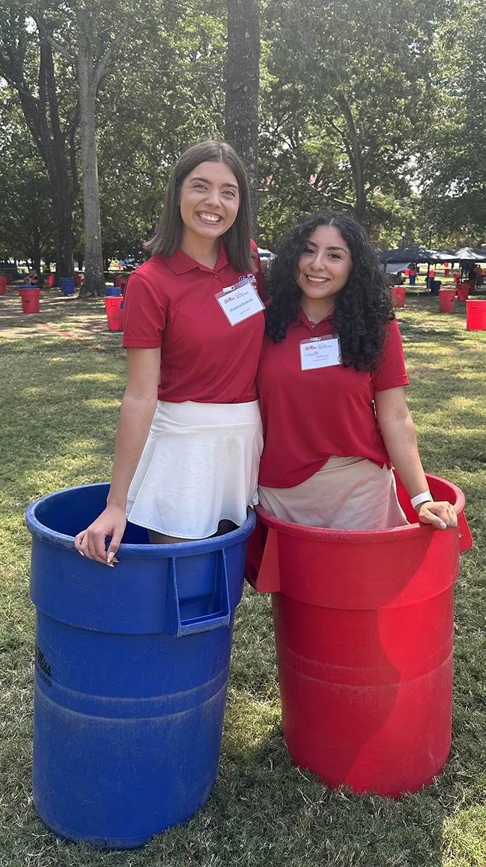 Two young women stand inside red and blue trash cans in the Grove
