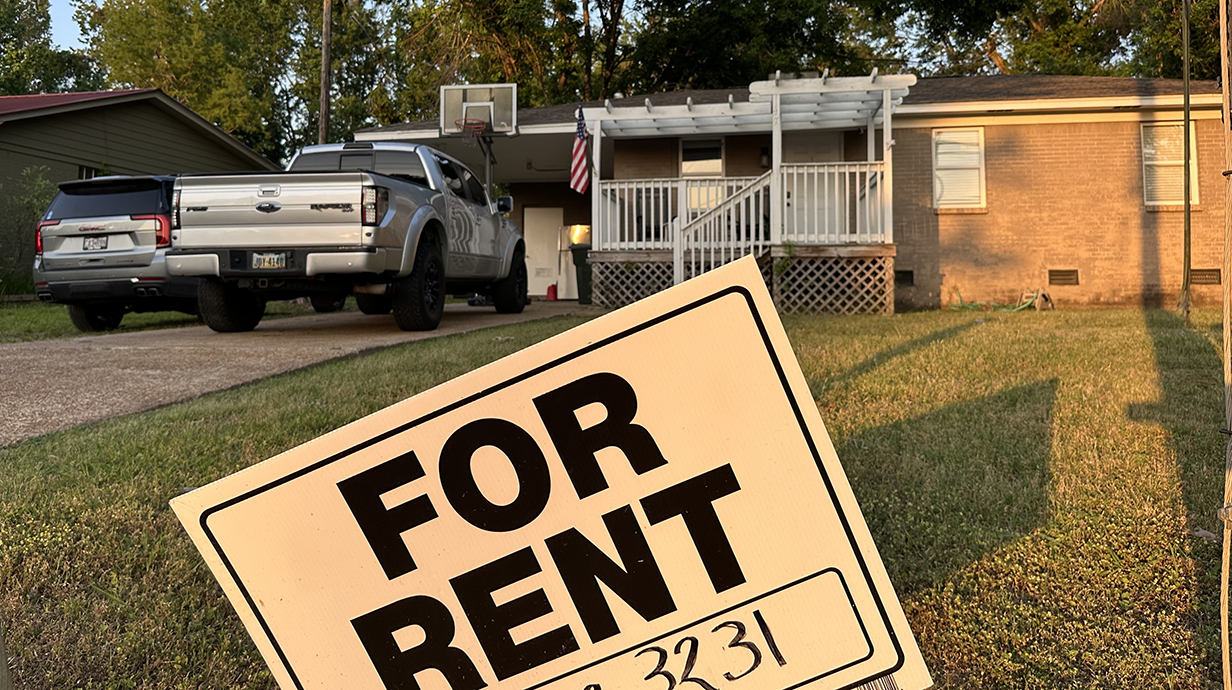 A 'For Rent' sign sits in front of a house with vehicles in the driveway.