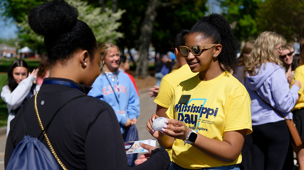 A young woman wearing a yellow T-shirt hands a stress ball to another young woman in an outdoor area.