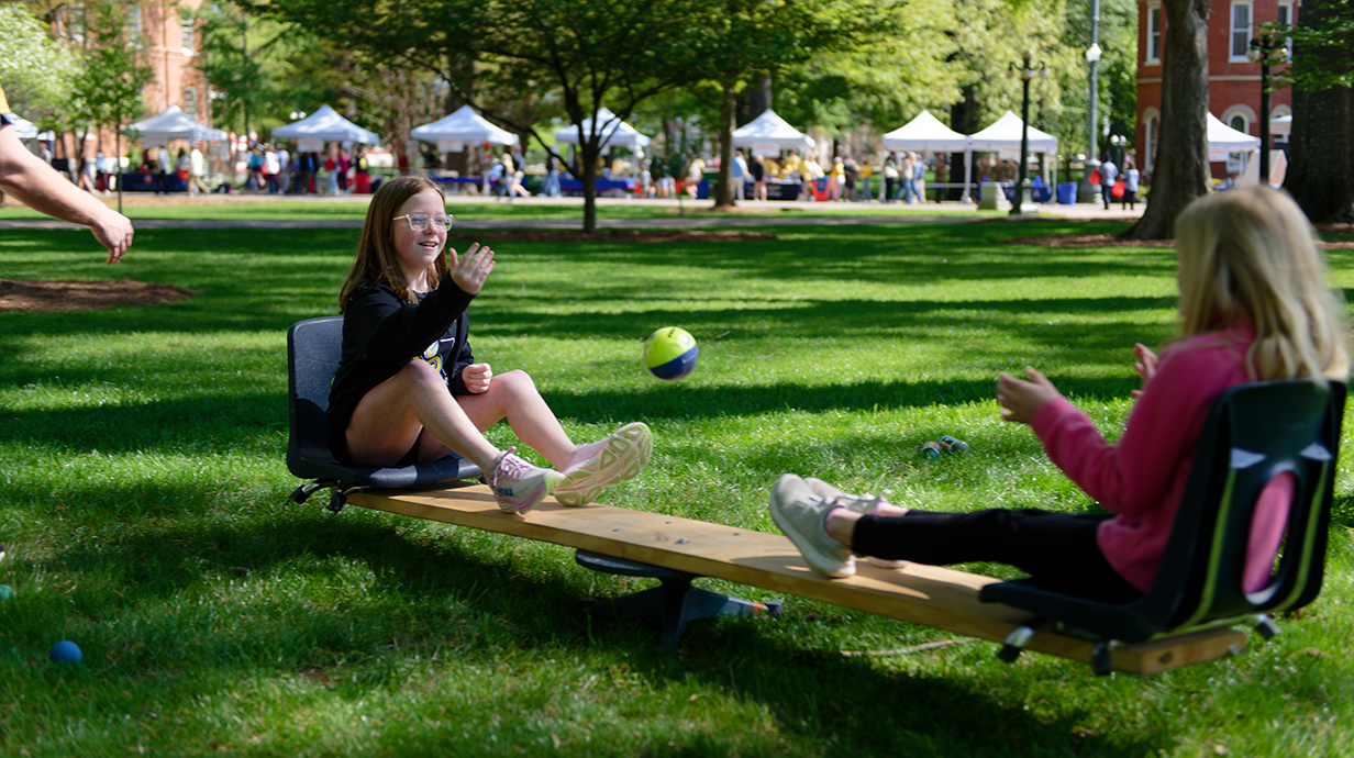 Two young girls sit on either end of a teeter-totter while tossing a ball between them.