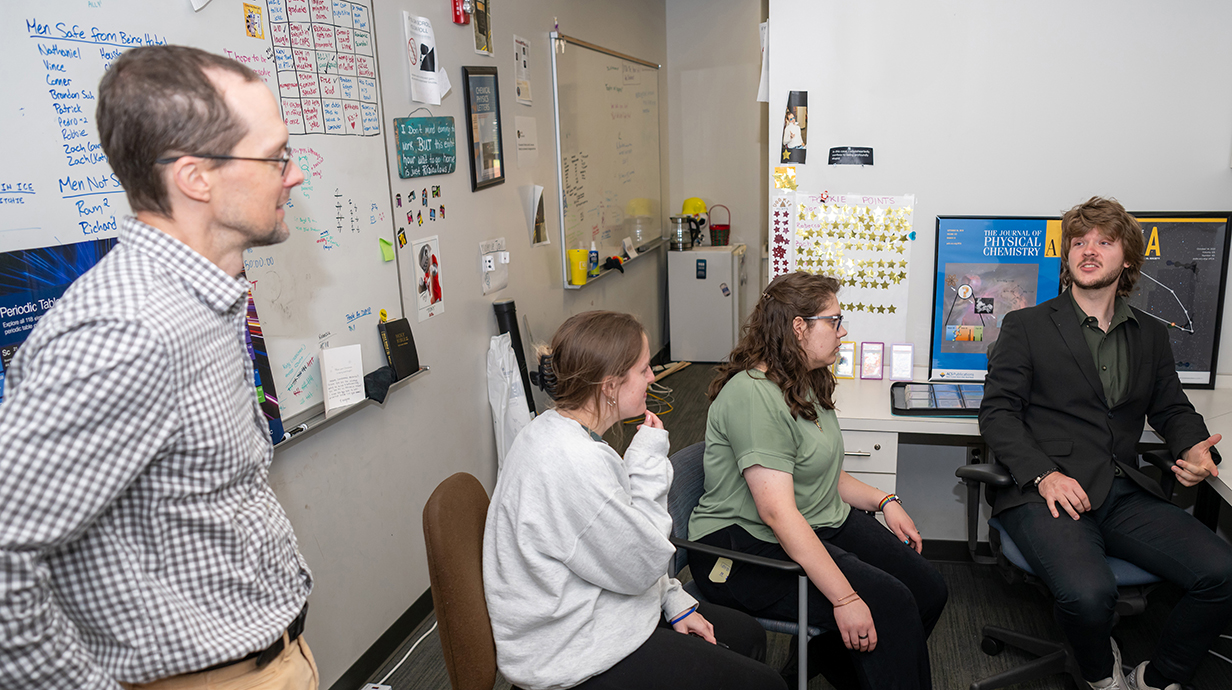 A young man seated at a computer talks to a man and two young women in a computer lab.