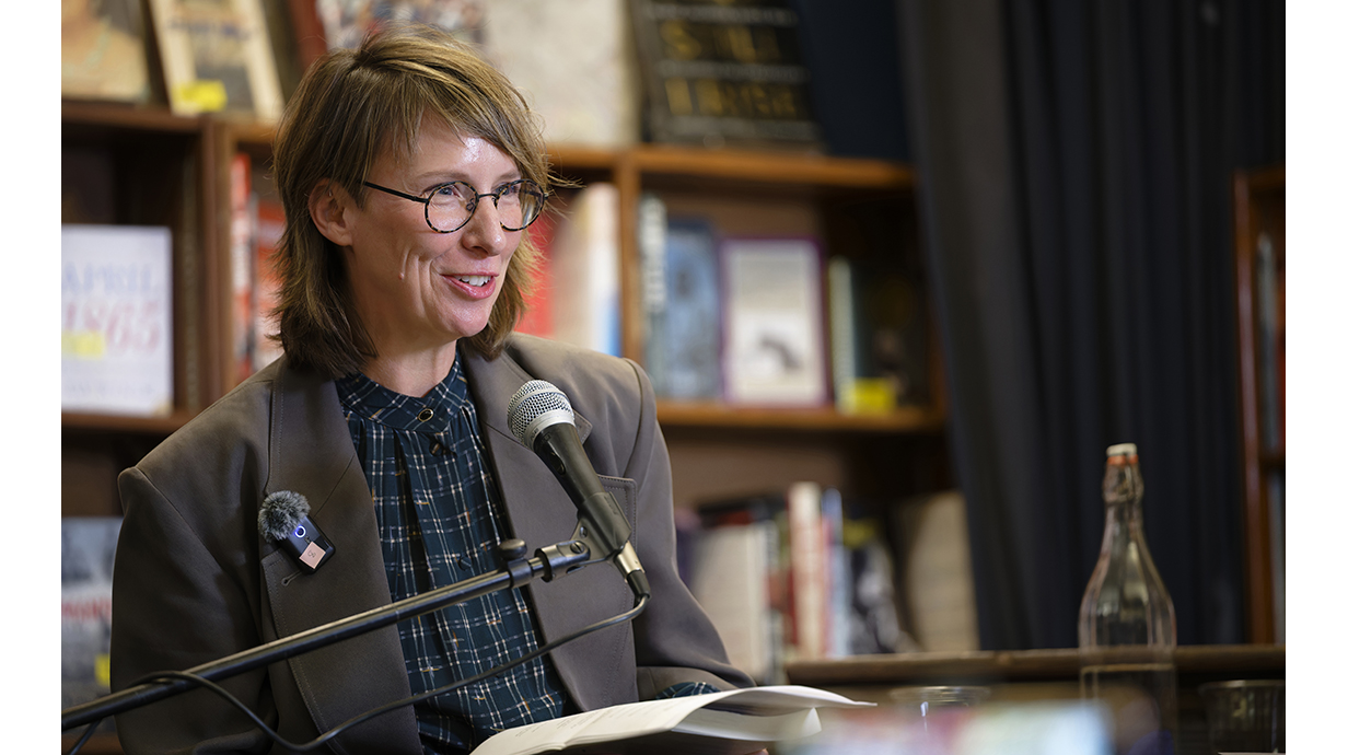 A woman holding a sheaf of papers and standing in front of bookcases speaks into a microphone.