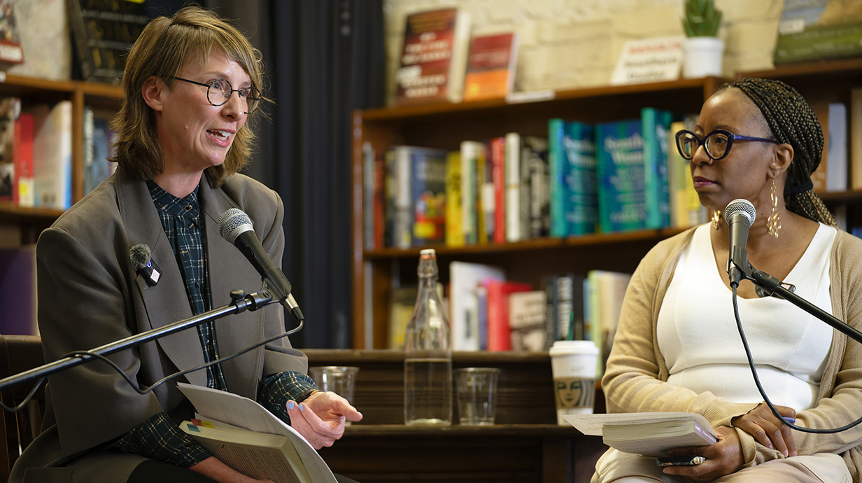 Two women seated in front of bookcases talk into stand microphones.