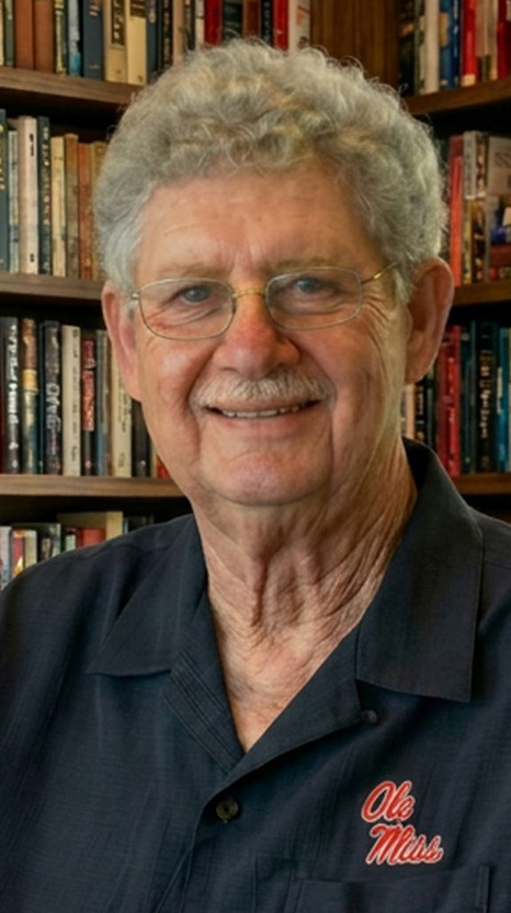 Headshot of a man wearing an Ole Miss polo shirt and sitting in front of bookcases filled with books.