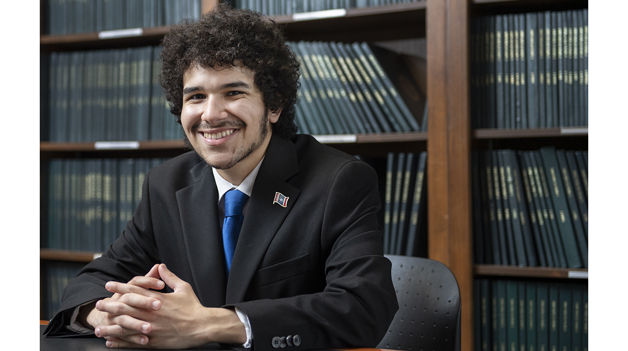 A young man wearing a suit sits at a conference table in front of bookshelves lined with law books.