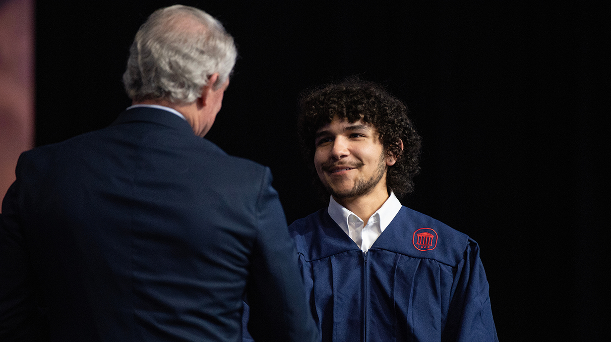 A man wearing a dark suit shakes hands with a young man wearing a graduation robe.
