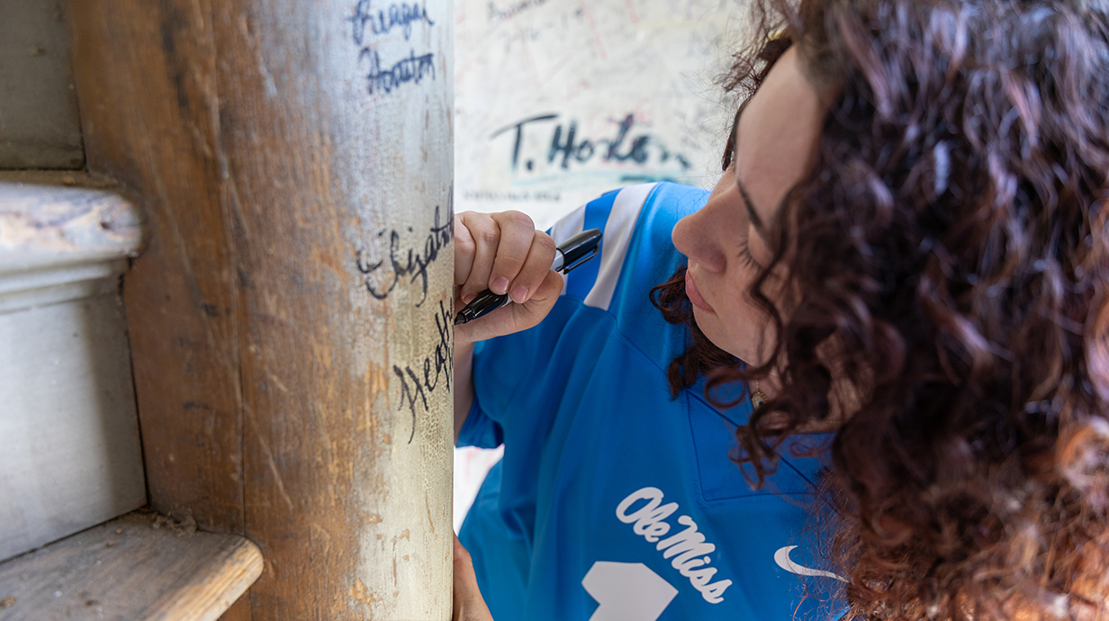 A young woman wearing a blue football jersey signs her name on a column in a building attic.