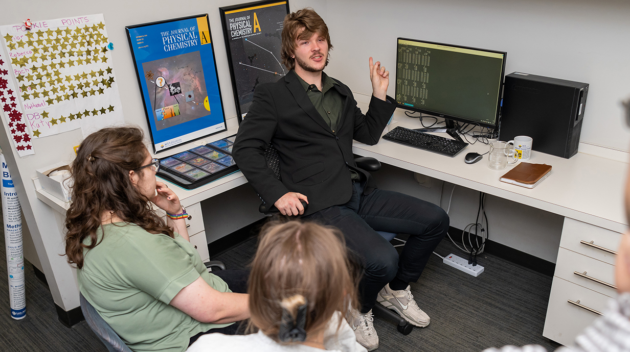 A young man wearing a casual black suit talks with a young man and young woman while seated in front of a computer.