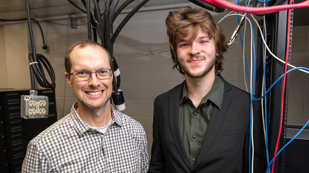 A man wearing glasses and a young man wearing a black casual suit stand in a lab filled with electrical cables and computer equipment.