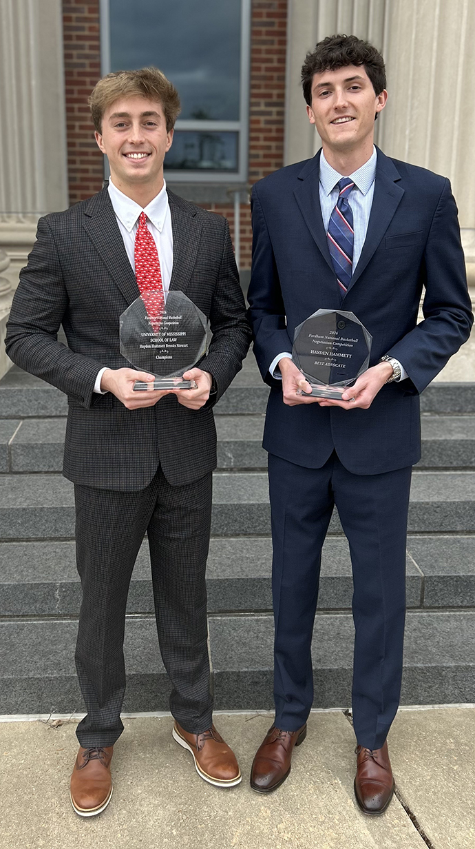 Two young men wearing suits, both holding crystal trophies, stand outside a large brick building.