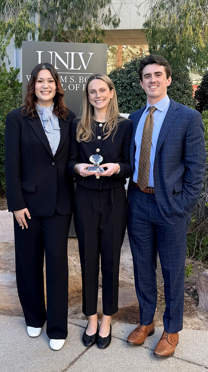 A young man and two young women, all dressed in professional attire, stand outdoors holding a small crystal trophy.