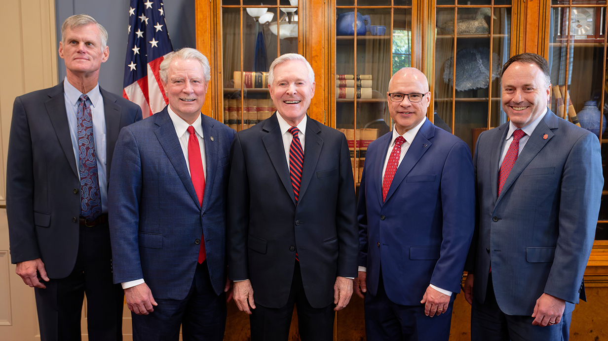 Five men, all wearing dark suits, stand in front of a glass-doored bookcase and an American flag in an ornate room.