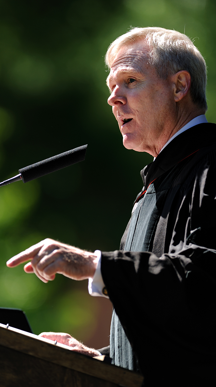 A man wearing a graduation robe speaks into a microphone at an outdoor podium.