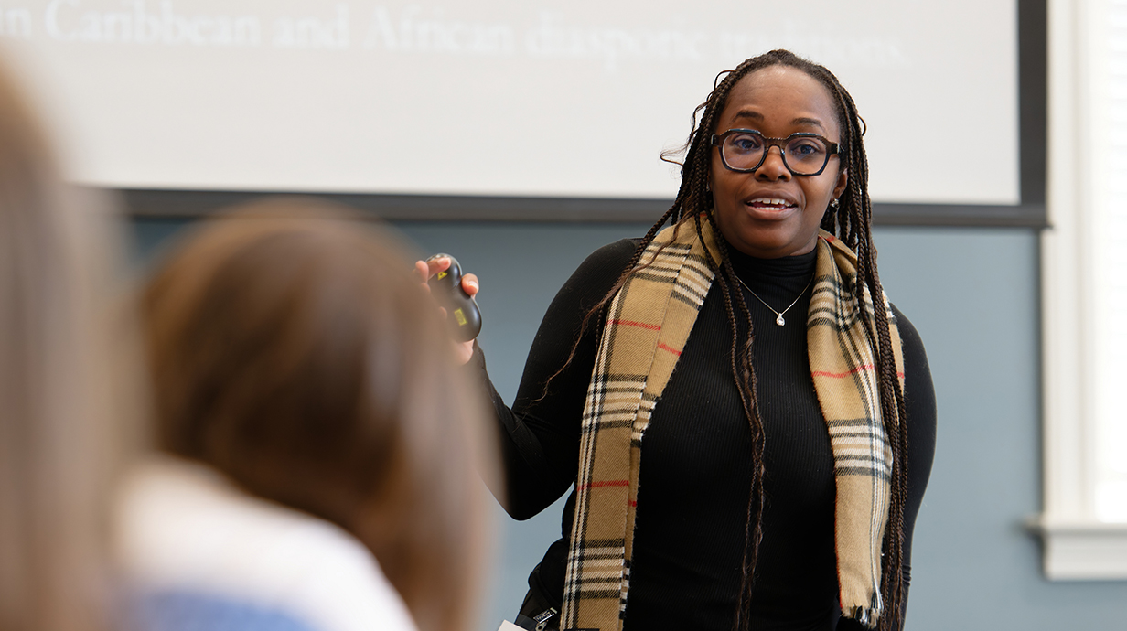 A woman wearing a tan plaid scarf over a black sweater talks to a classroom of students.