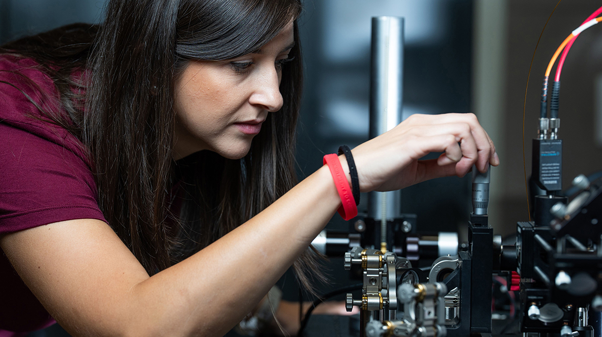 A woman wearing a crimson shirt adjusts a piece of equipment in a lab.