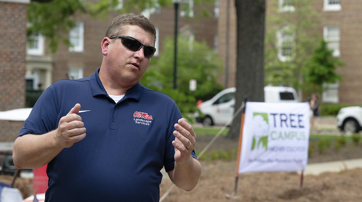 A man gestures as he talks in front of a banner reading 'Tree Campus.'