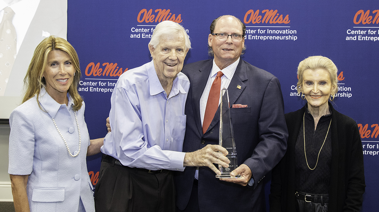 Two women and two men, one holding a crystal trophy, stand in front of a dark blue backdrop covered with Ole Miss logos.