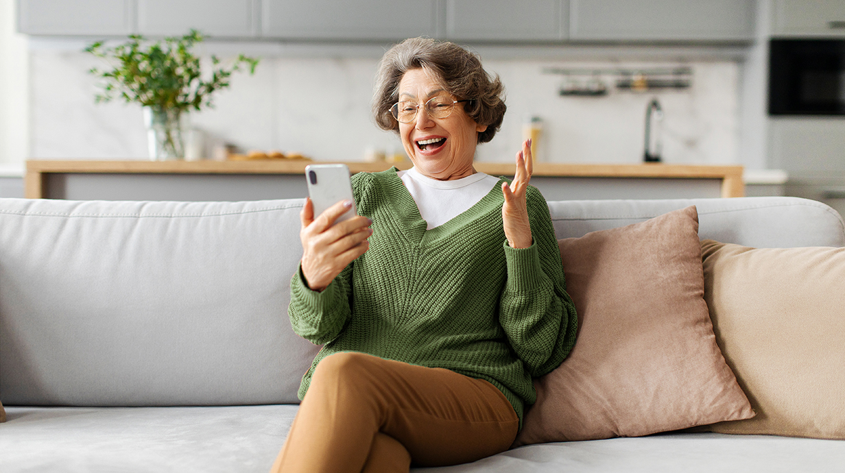 An older woman sitting on a sofa throws up her hands in delight as she reads something on a cellphone.