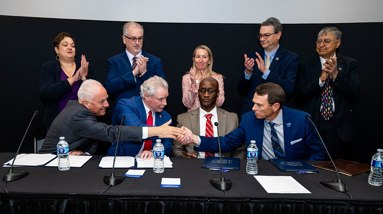 Four men sitting a a table covered with a black drape shake hands as  three men and two women standing behind them applaud.