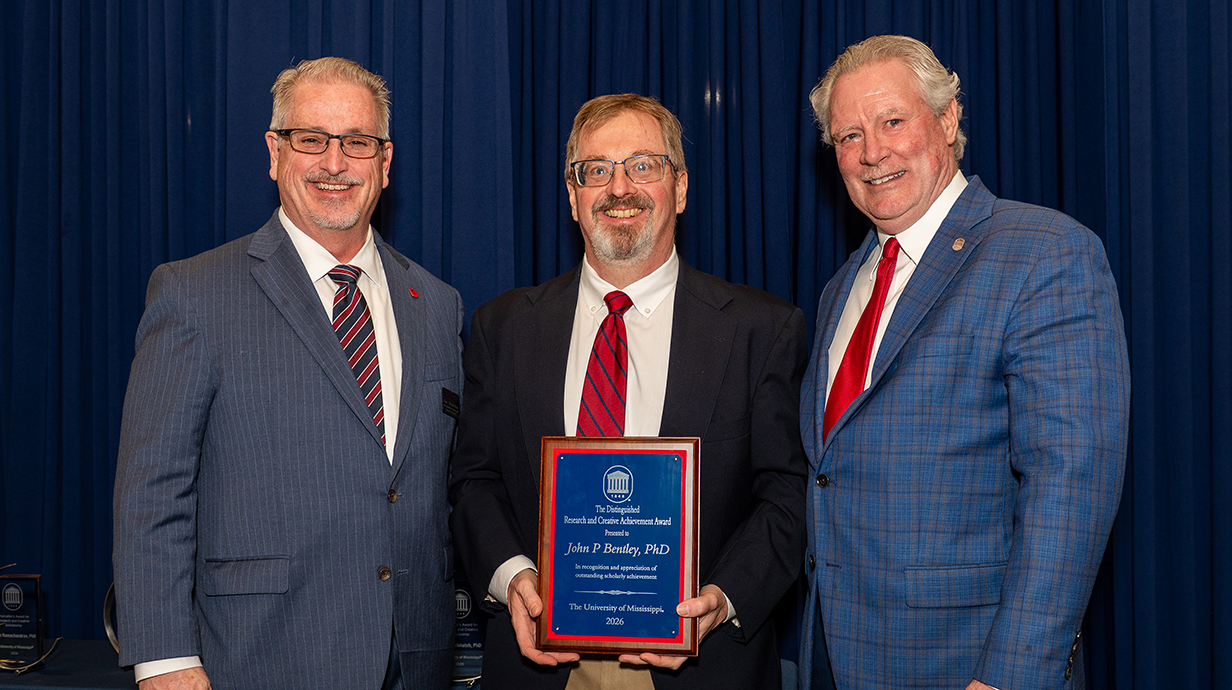 Three men wearing suits, the middle one holding an award plaque, stand in front of a blue backdrop.