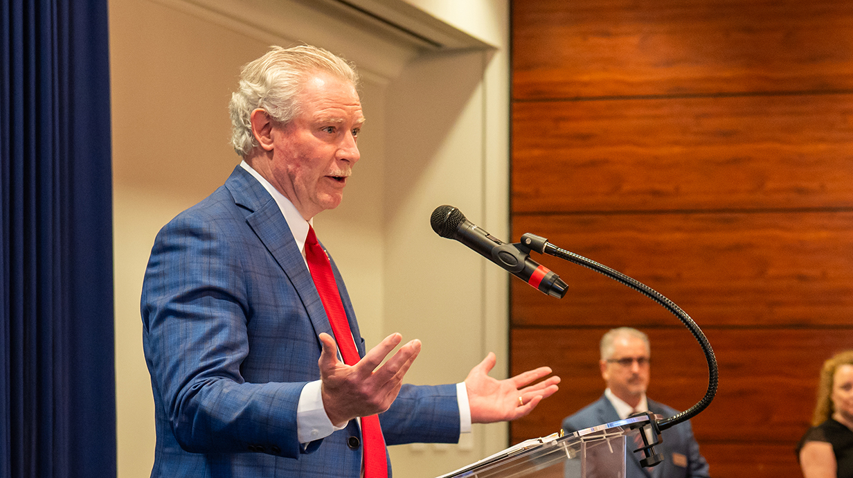 A man wearing a suit speaks into a microphone at the front of a banquet room.