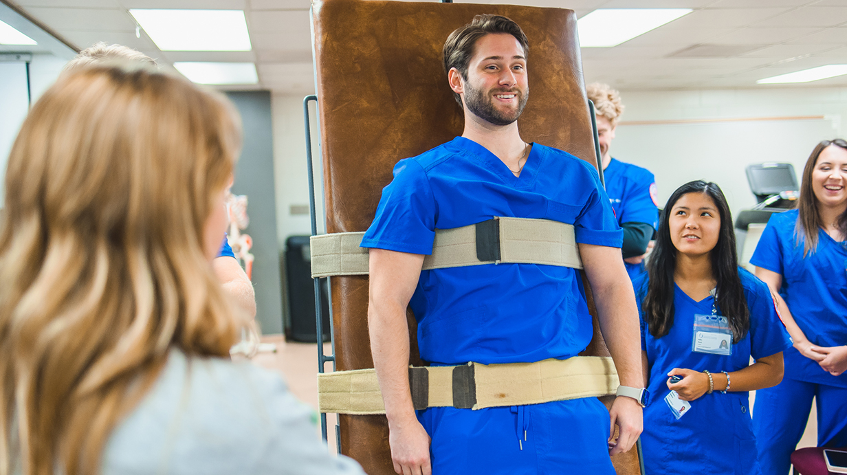 A young man wearing blue medical scrubs stands strapped to a table while other people wearing scrubs watch.