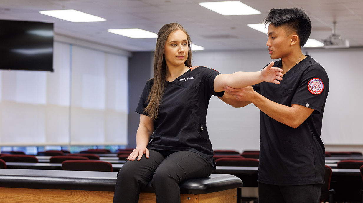 A young man wearing black medical scrubs hold the outstretched arm of a young woman, also wearing scrubs.